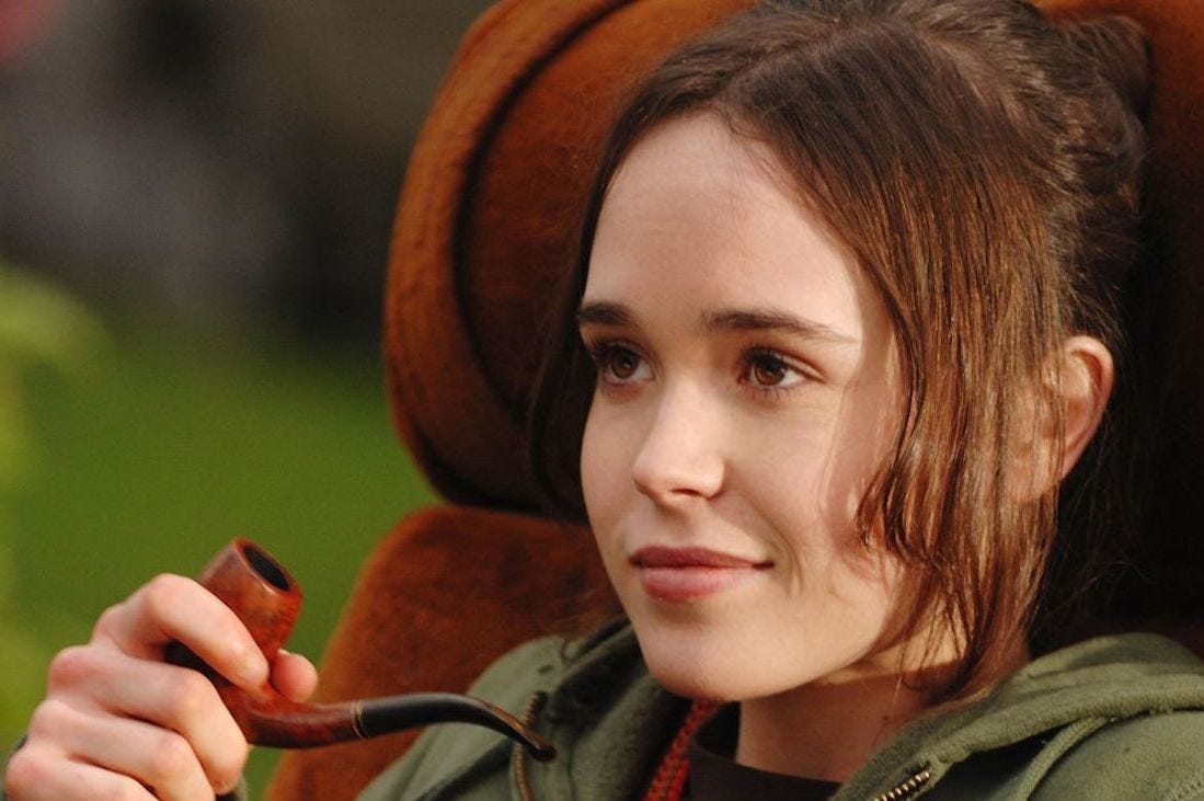 Elliot Page as Juno, smirking while sat in a brown chair and holding a pipe. 