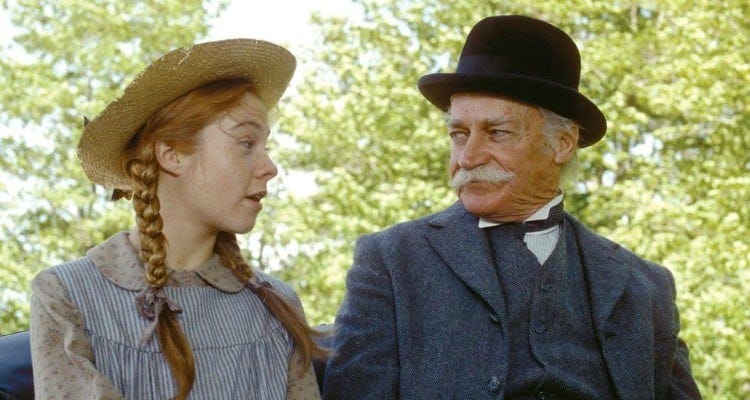 Megan Follows in her straw hat and pigtails, mid-sentence, speaks to a bemused, mustachioed Matthew in a dark gray suit and black bowler hat. Behind them are sunlit trees.