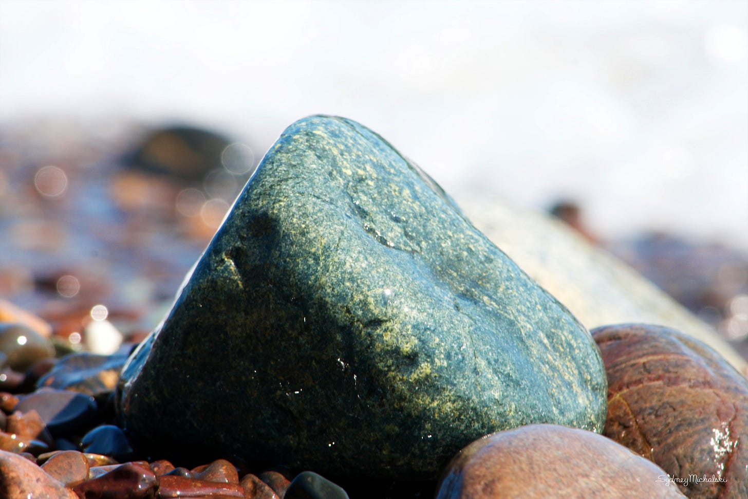A greed and gold stone shimmers in the salt spray and sunshine of a cobble beach.