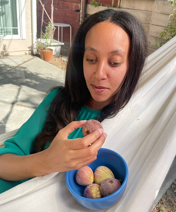 I’m a Black woman with long dark hair holding a fig. I’m outside in a hammock, and the fabric is partially folded to create a shelf for a bowl with large, red & green figs.