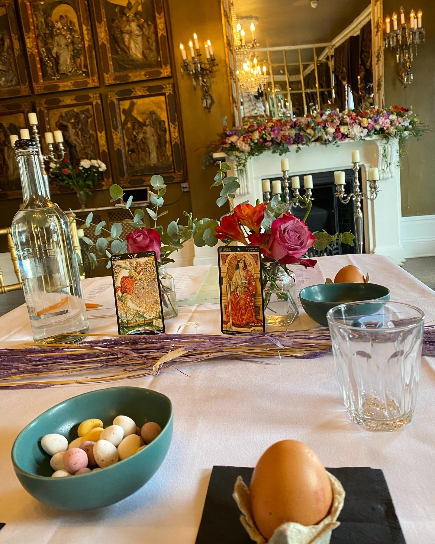 A table with flowers, an egg, and tarot cards in a grand room with wall chandeliers 