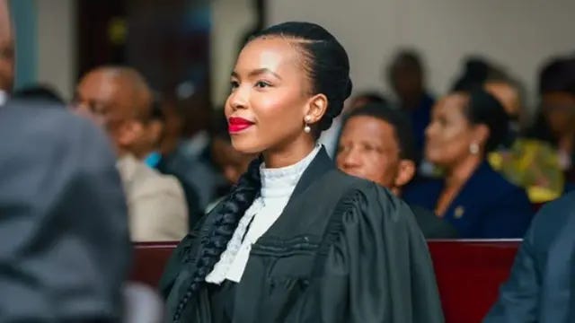 Lesego Chombo in a black legal gown and white collar. Wearing red lipstick, she is seated on a bench with others in a courtroom and  is looking to the side - not at the camera.
