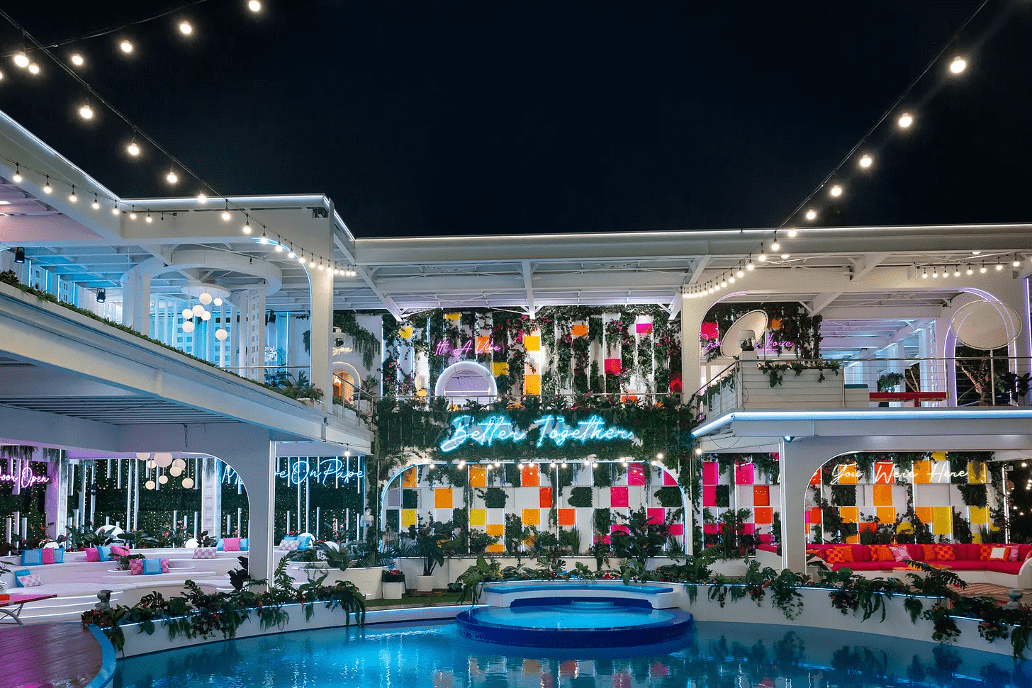 A multi-level villa featuring a central swimming pool, white modern architecture with string lighting, vibrant colored wall panels, a teal neon "Better Together" sign, and resort-style lounge areas with tropical plants creating an upscale social atmosphere. A multi-level villa featuring a central swimming pool, white modern architecture with string lighting, vibrant colored wall panels, a teal neon "Better Together" sign, and resort-style lounge areas with tropical plants creating an upscale social atmosphere.