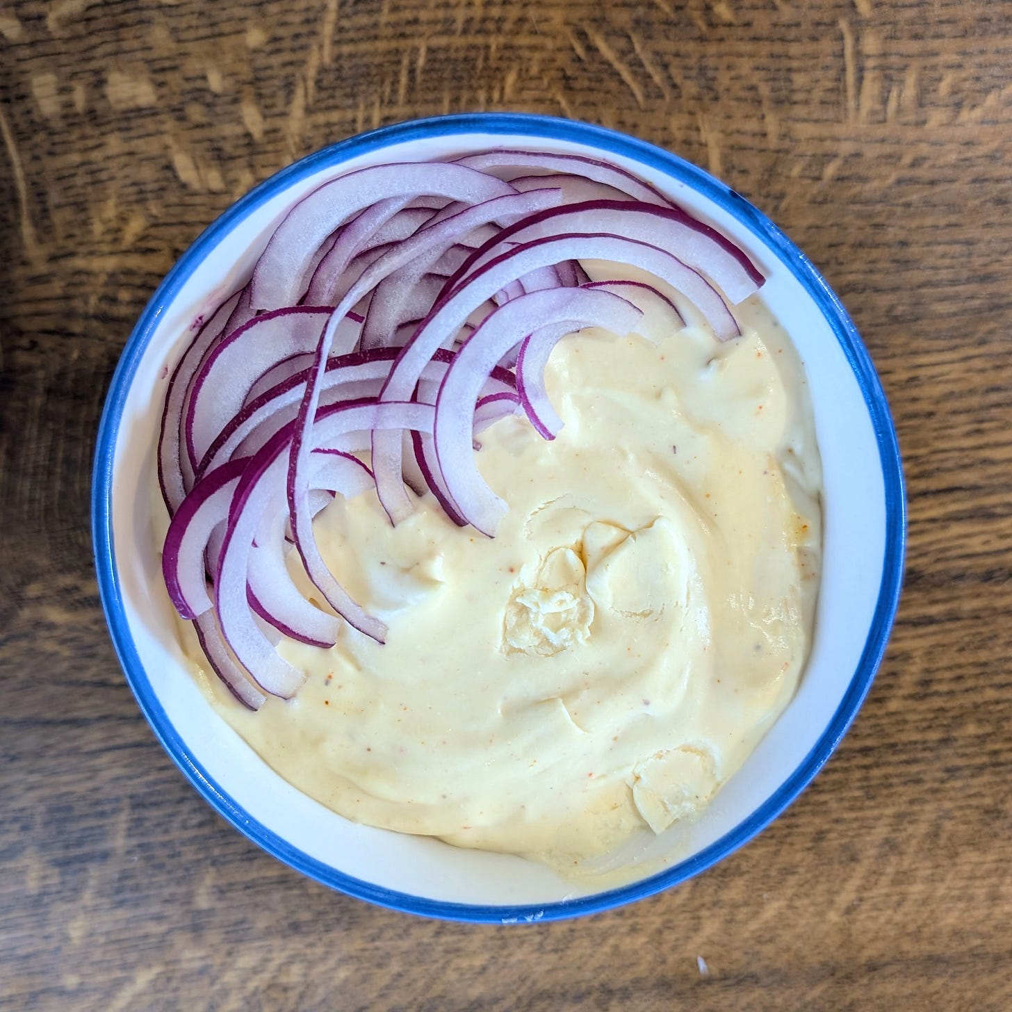 A small bowl with a blue‑rimmed edge holds a creamy pale yellow spread topped with thin semicircles of red onion. The bowl sits on a wooden surface, highlighting the contrast between the smooth dip and the crisp onion slices.