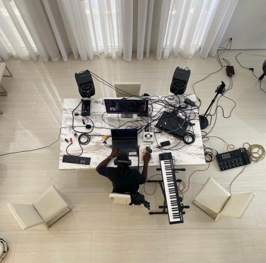 A black person at a white table in a white room surrounded by electronics. A black person at a white table in a white room surrounded by electronics.
