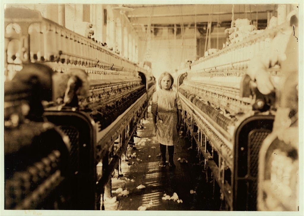 Old black and white photo of a young girl working in a textile mill. She is surrounded on both sides by hundreds of spools and bobbins.