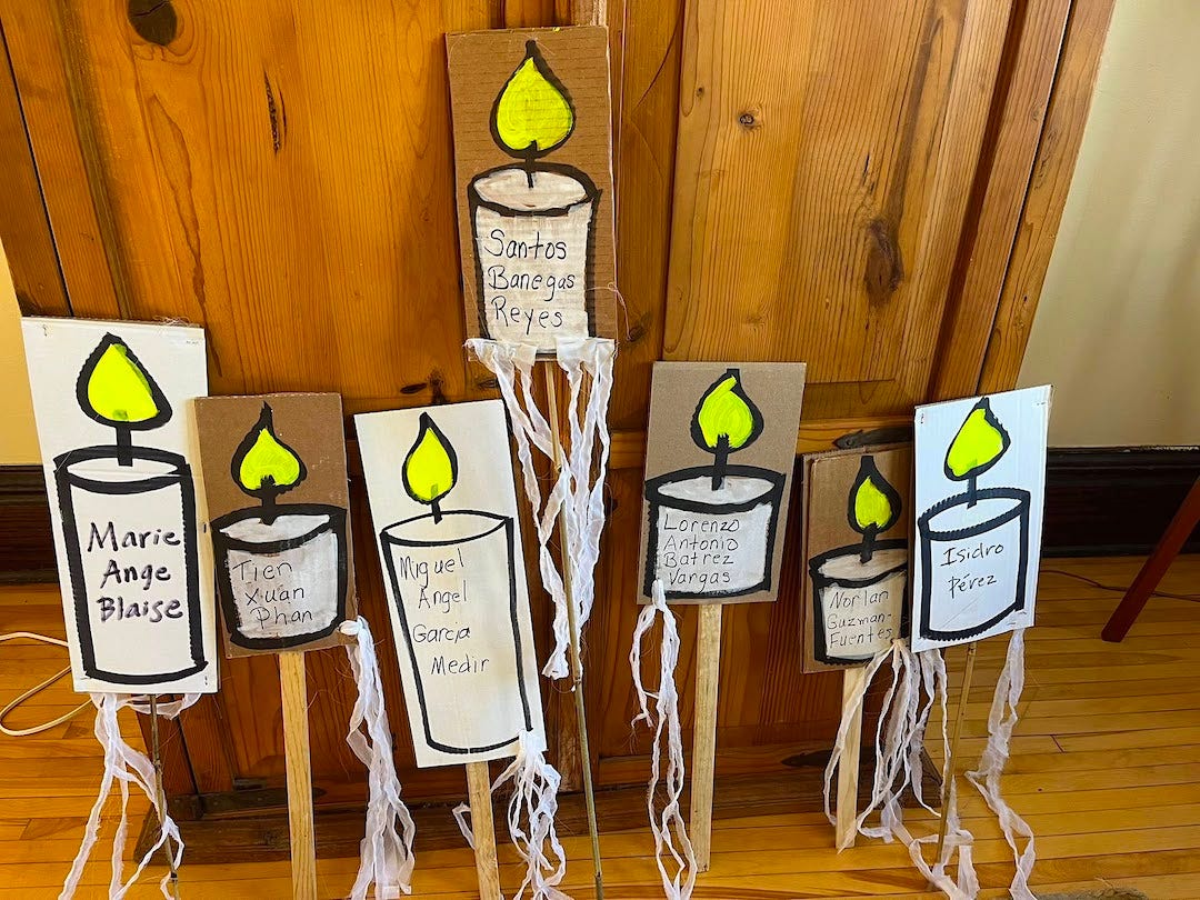 Seven hand painted signs lean against a wooden door. Each depicts a burning candle with the name of somebody killed by ICE. Long white raffia tassles hang from each, like flickering light.