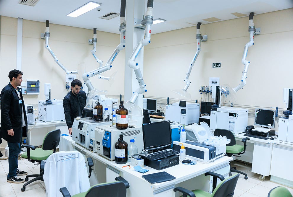 Laboratório científico com duas pessoas em pé, uma à esquerda usando jaqueta preta e outra ao centro também em jaqueta preta, ambas próximas a bancadas brancas equipadas com computadores portáteis, garrafas de vidro âmbar, equipamentos de análise e instrumentos de laboratório, com múltiplos braços articulados brancos com pontos azuis suspensos do teto, cadeiras com assento verde e encosto preto distribuídas pelo espaço, paredes bege claro, iluminação fluorescente no teto, e diversos equipamentos eletrônicos brancos e pretos organizados nas superfícies de trabalho. Laboratório científico com duas pessoas em pé, uma à esquerda usando jaqueta preta e outra ao centro também em jaqueta preta, ambas próximas a bancadas brancas equipadas com computadores portáteis, garrafas de vidro âmbar, equipamentos de análise e instrumentos de laboratório, com múltiplos braços articulados brancos com pontos azuis suspensos do teto, cadeiras com assento verde e encosto preto distribuídas pelo espaço, paredes bege claro, iluminação fluorescente no teto, e diversos equipamentos eletrônicos brancos e pretos organizados nas superfícies de trabalho.