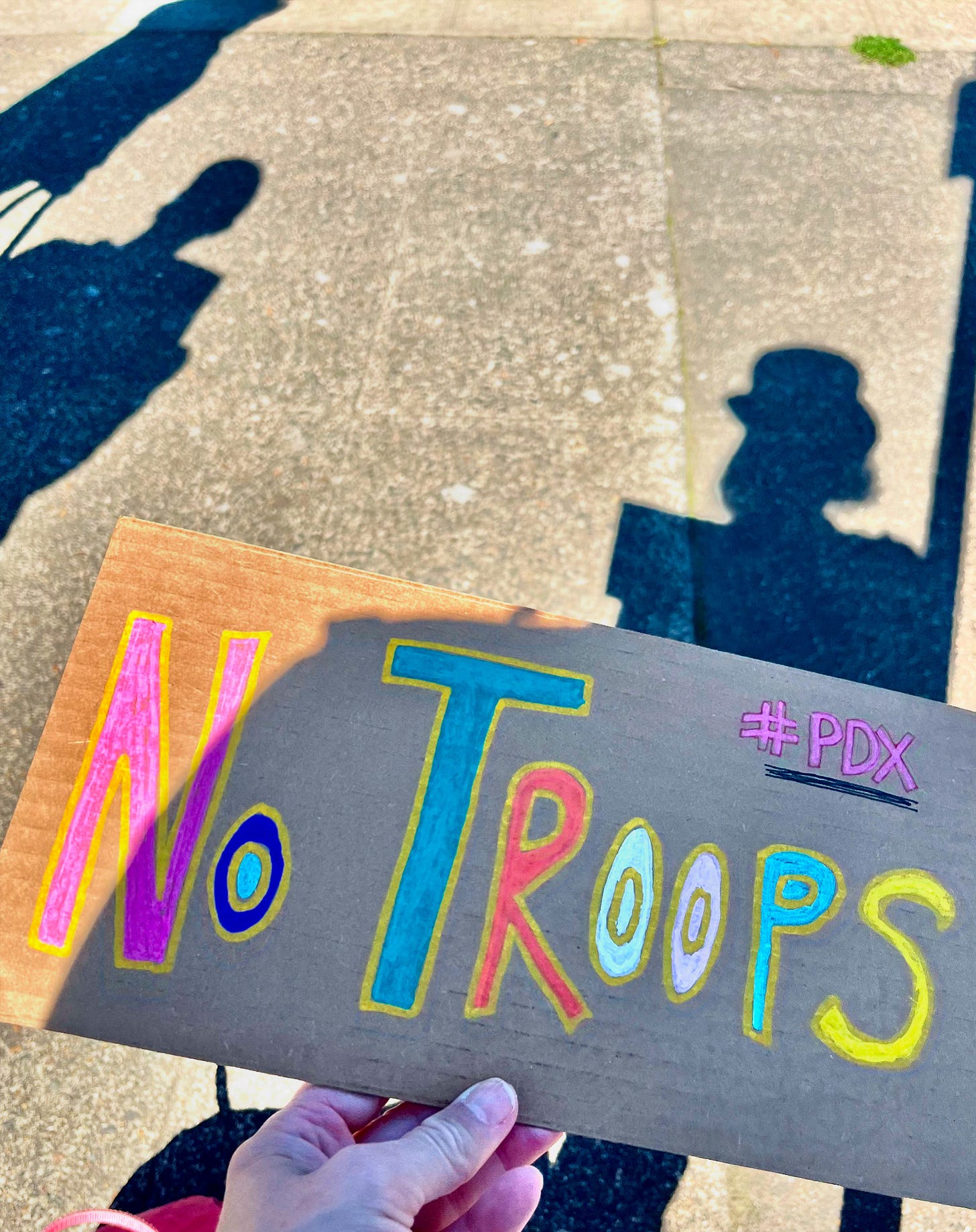 Shadows of people; in the foreground NO TROOPS is on a cardboard sign, written in bright paint pens.