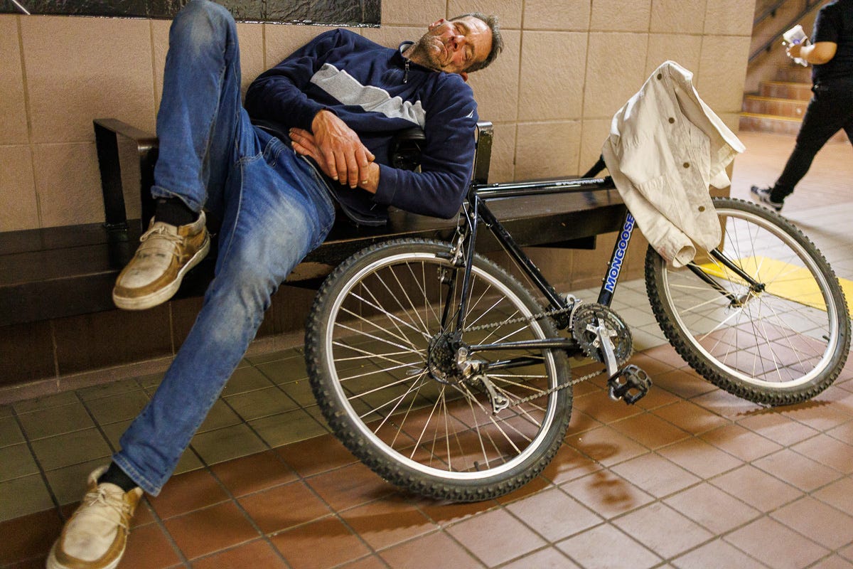 A man in jeans and a dark pullover sleeping on a subway station bench with a Mongoose bicycle leaned beside him and a white jacket hanging over the handlebars. A man in jeans and a dark pullover sleeping on a subway station bench with a Mongoose bicycle leaned beside him and a white jacket hanging over the handlebars.
