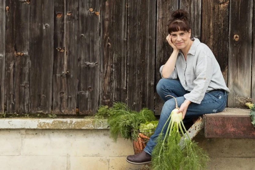 Die Autorin Esther Kern sitzt entspannt auf einer niedrigen Rampe vor einer dunklen, verwitterten Holzwand. Sie trägt ein helles Shirt, blaue Jeans und braune Schuhe, stützt den Kopf in die Hand und hält einen frischen Fenchel mit langen, grünen Blattfedern. Neben ihr steht ein Korb voller Gemüse.