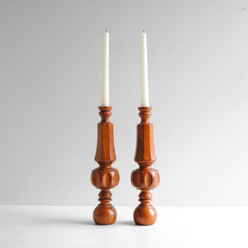 May include: Two wooden candle holders with white candles. The candle holders are a warm brown color with a turned design. The base is round, and the body has a series of rounded sections. The candles are tall and slender.