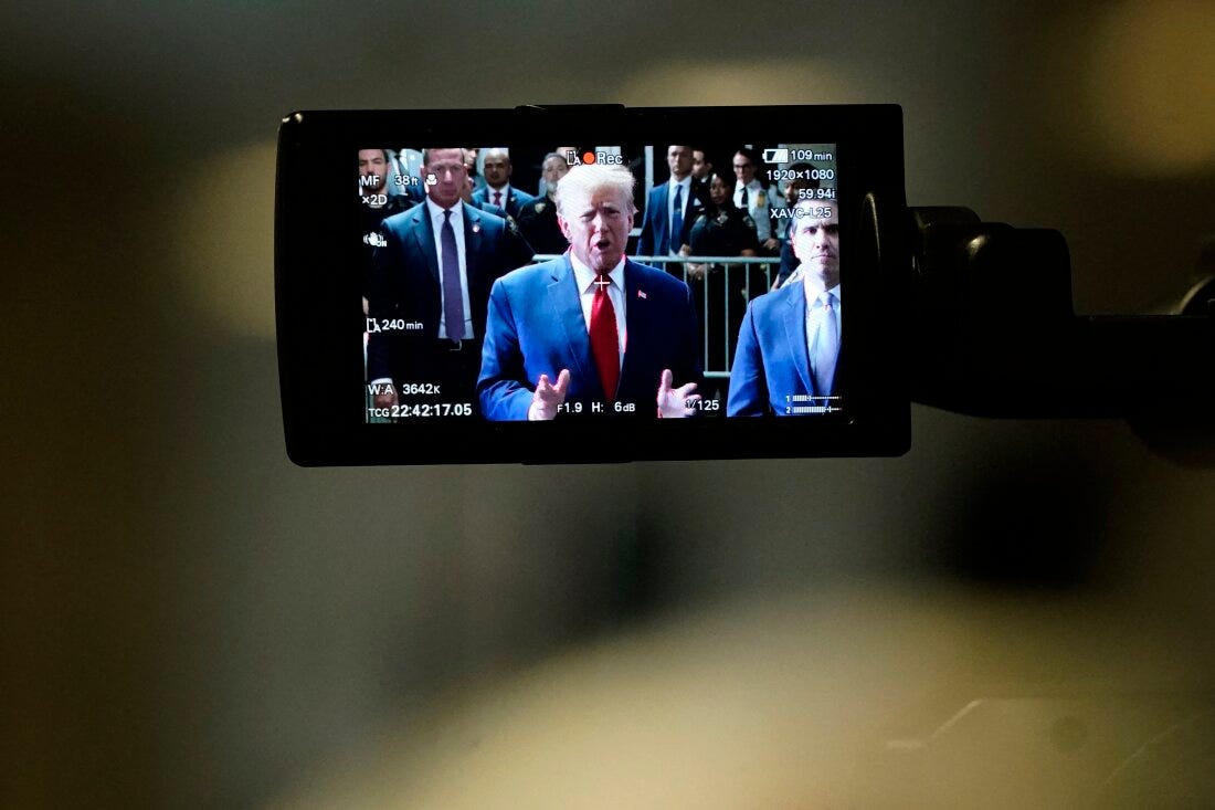 This photo shows former President Donald Trump on a camera monitor as he speaks to the press in February outside Manhattan Criminal Court. He's wearing a blue suit jacket, a red tie and a white shirt. Many people in suits and law enforcement uniforms stand behind him. 