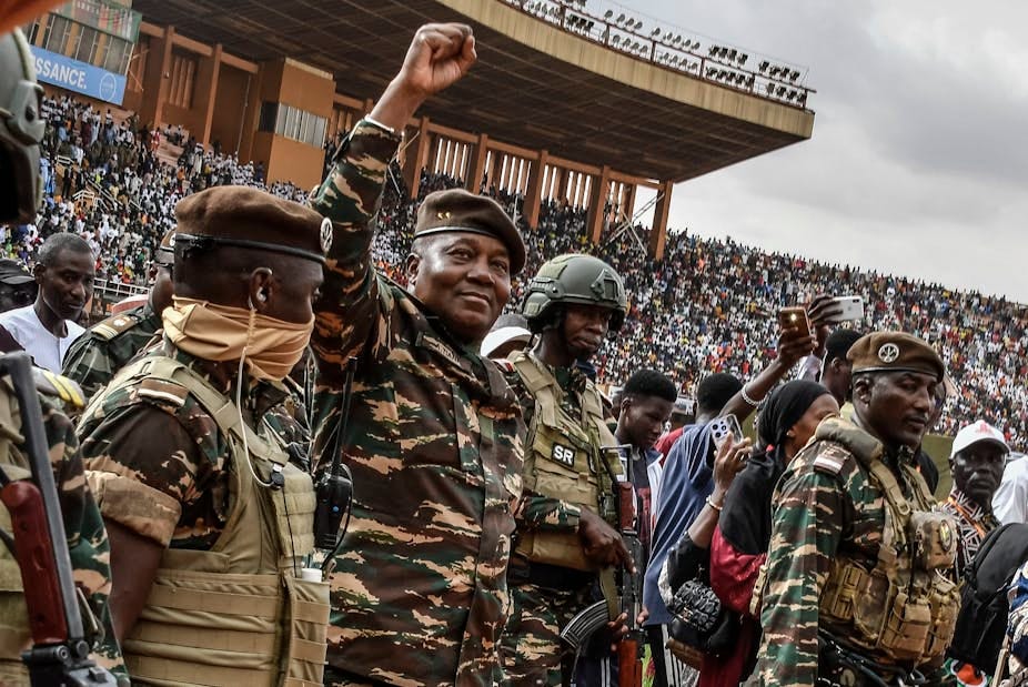 A smiling man dressed in army fatigues holding up a fisted arm while surrounded by armed military men, with a crowd of seated people behind him in a stadium A smiling man dressed in army fatigues holding up a fisted arm while surrounded by armed military men, with a crowd of seated people behind him in a stadium