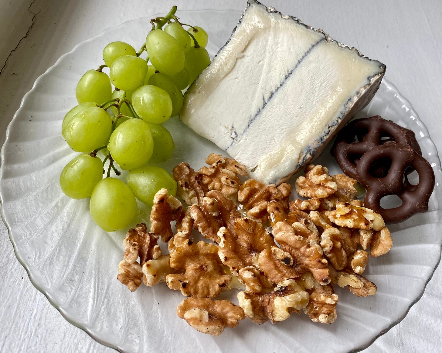 Humboldt Fog slice on a clear plate next to chocolate covered pretzels, toasted walnuts with honey, and green grapes