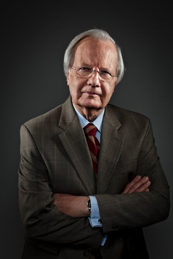 Wearing a suit and tie, he stands with his arms crossed and looks directly into the camera. He has gray hair and glasses.