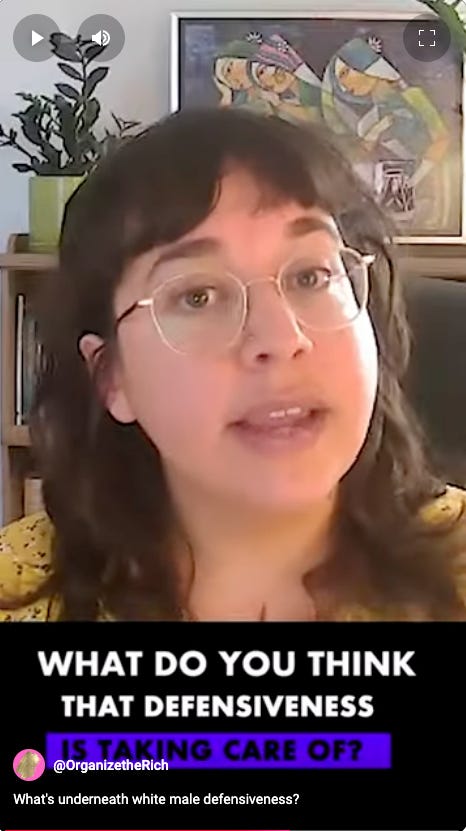 Screenshot photo from the video clip. Iris is in the full frame, looking at the camera and talking. In a yellow shirt. Art on the walls behind her. The caption reads "What do you think the defensiveness is taking care of?"