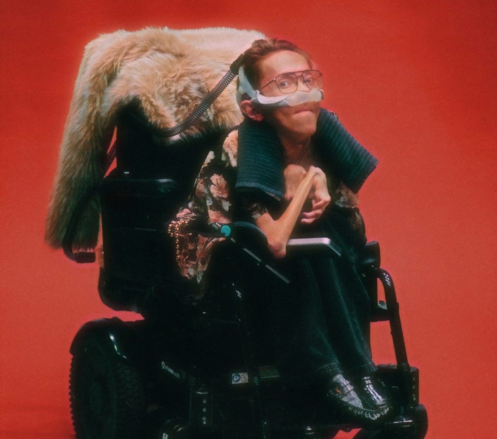 A glossy portrait with a soft 80s glow. A white disabled man with a nasal ventilator mask, towel around his neck, and a fur coat hanging off the back of his powerchair.