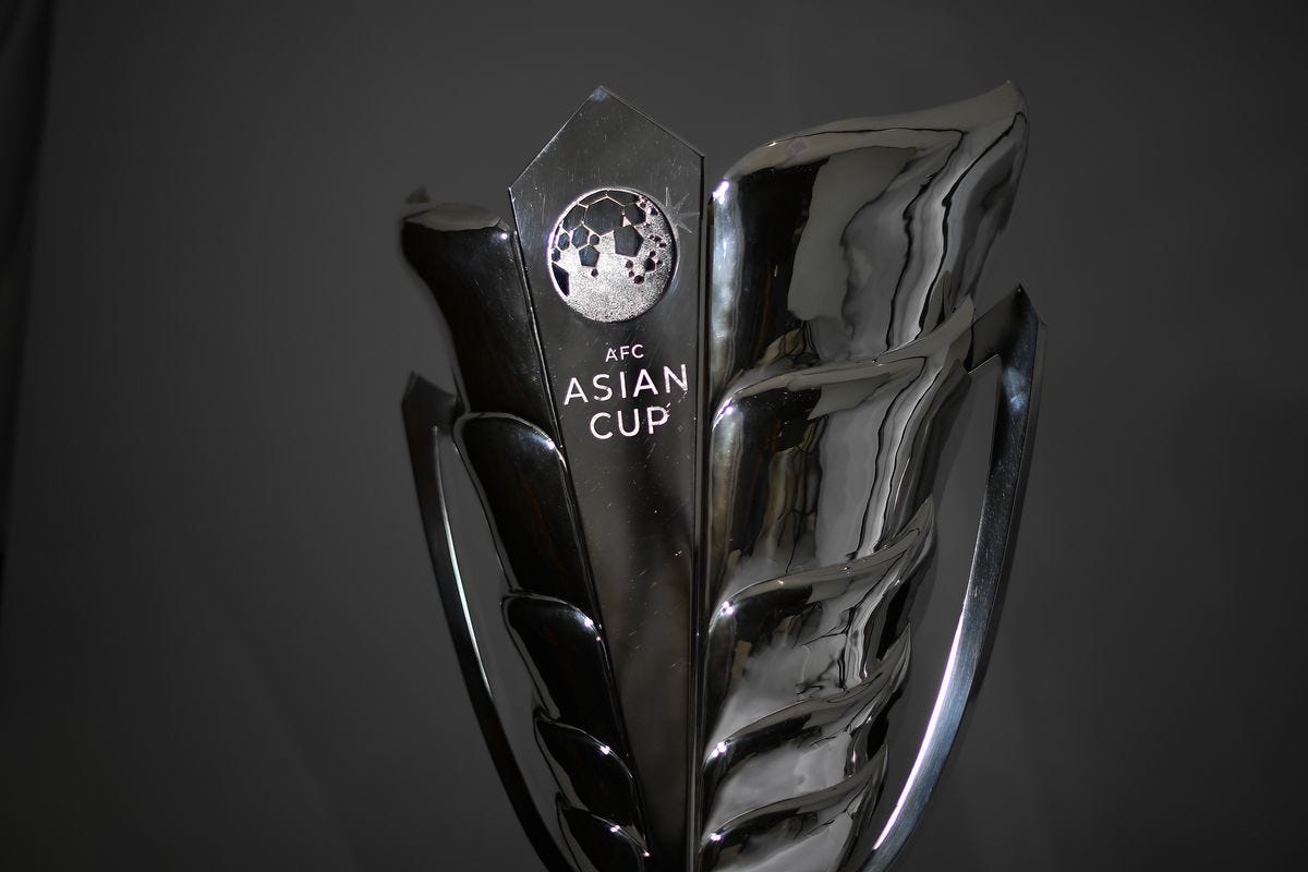 A close-up photograph of the ornate, highly polished silver AFC Asian Cup trophy against a plain dark gray background. The central panel displays a textured AFC logo with a star pattern and the words ‘AFC ASIAN CUP’ in two lines.