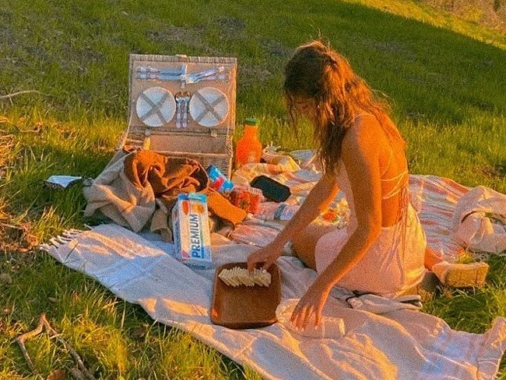 This may contain: a woman sitting on a blanket in the grass next to an open suitcase with food
