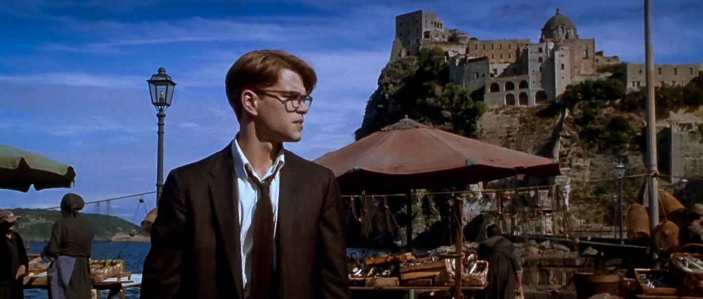 Matt Damon as Tom Ripley in a dark suit and tie, standing in a seaside Italian market with Castello Aragonese in the background. His face is in partial shadow as he looks left, detached from the lively world behind him. Matt Damon as Tom Ripley in a dark suit and tie, standing in a seaside Italian market with Castello Aragonese in the background. His face is in partial shadow as he looks left, detached from the lively world behind him.