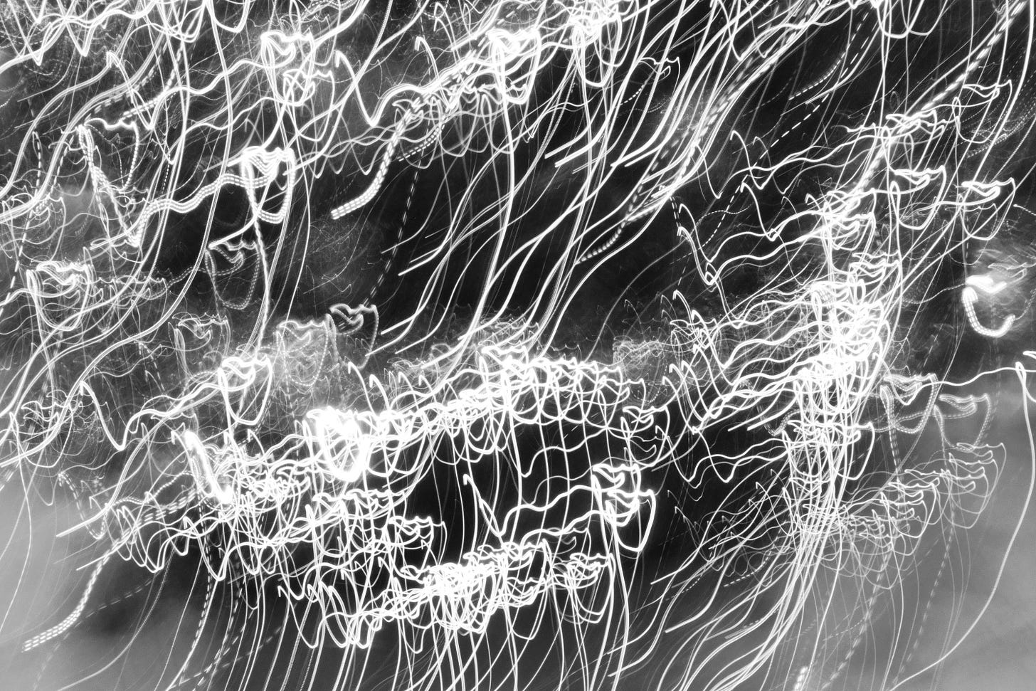 Black-and-white abstract photograph of bright, continuous light trails. The lines twist, loop, and overlap chaotically across the frame, creating a dense web of glowing streaks. The effect resembles scribbled calligraphy or tangled threads of light, with areas of both tight clusters and open flowing arcs.