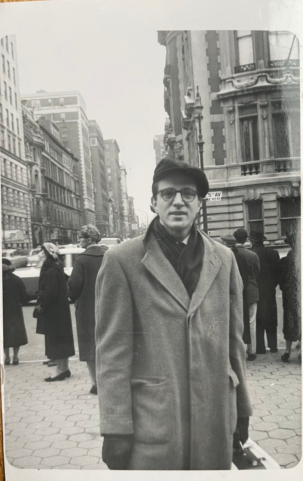 A man stands on a busy street corner, buildings and people behind him; he wears glasses, a cap and overcoat, and holds the handle of a case of some kind. He wears a tie under a scarf, but his expression is warm and informal.