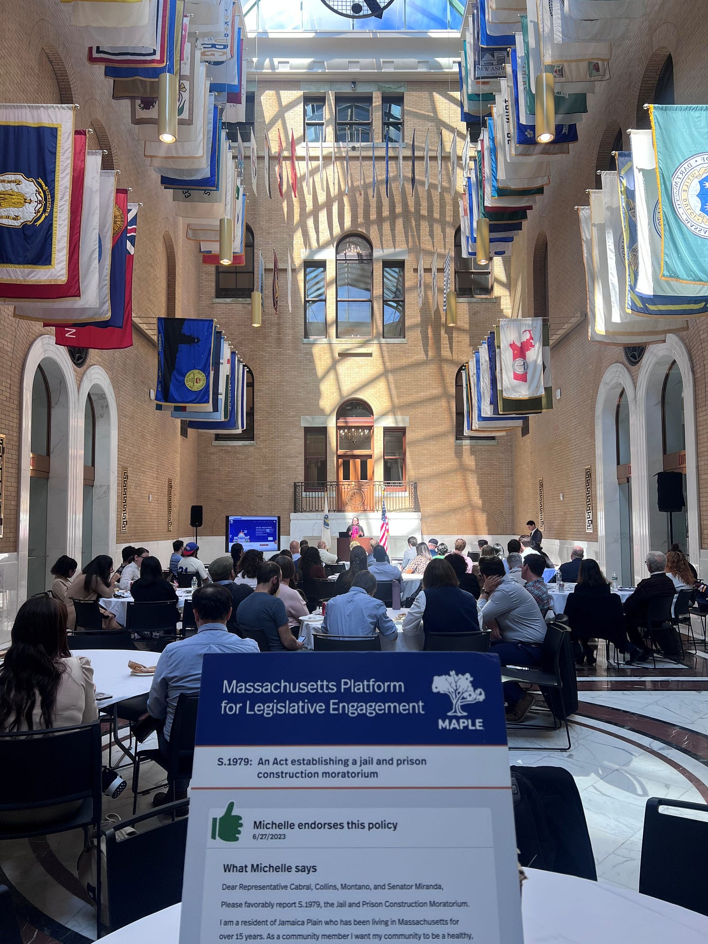 an audience sits in a large atrium facing a speaker at a podium with two flags in the background. in the foreground, a sign with MAPLE's name and a comment by a constituent on a policy about a prison construction moratorium