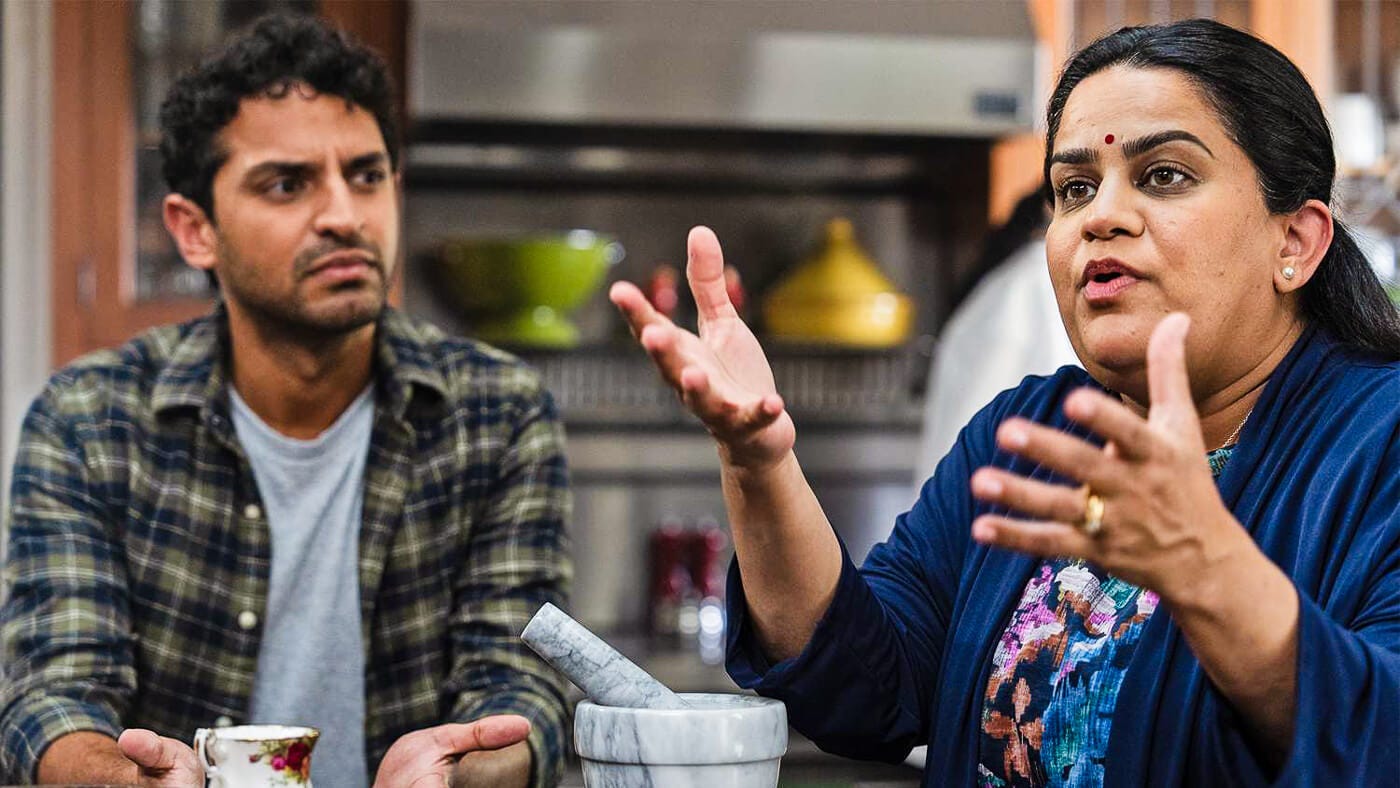 Megha speaks passionately in the kitchen with her hands raised as Naveen sits beside her, reacting with concern and confusion.