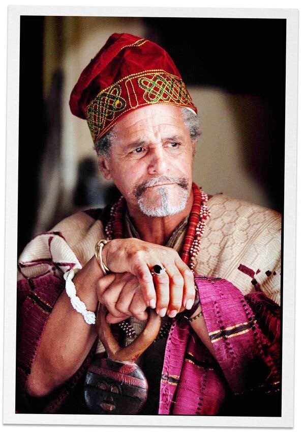 Adefunmi I as an older man with a graying beard in his African garb, complete with a red cap. He is looking off to the side with a pensive expression.