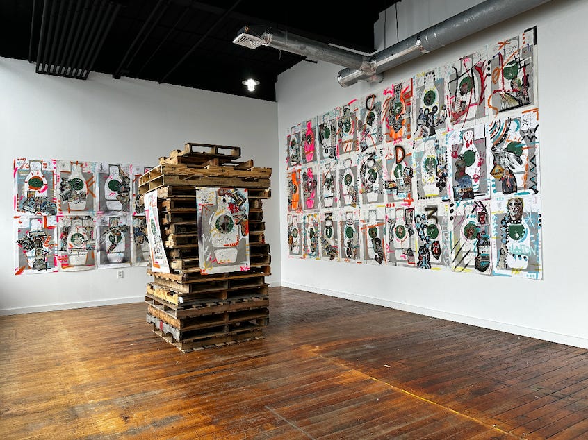 Open gallery space with wooden floors and white walls. A stack of wooden palettes is in the center of the room and there is artwork on the palettes and surrounding gallery walls.