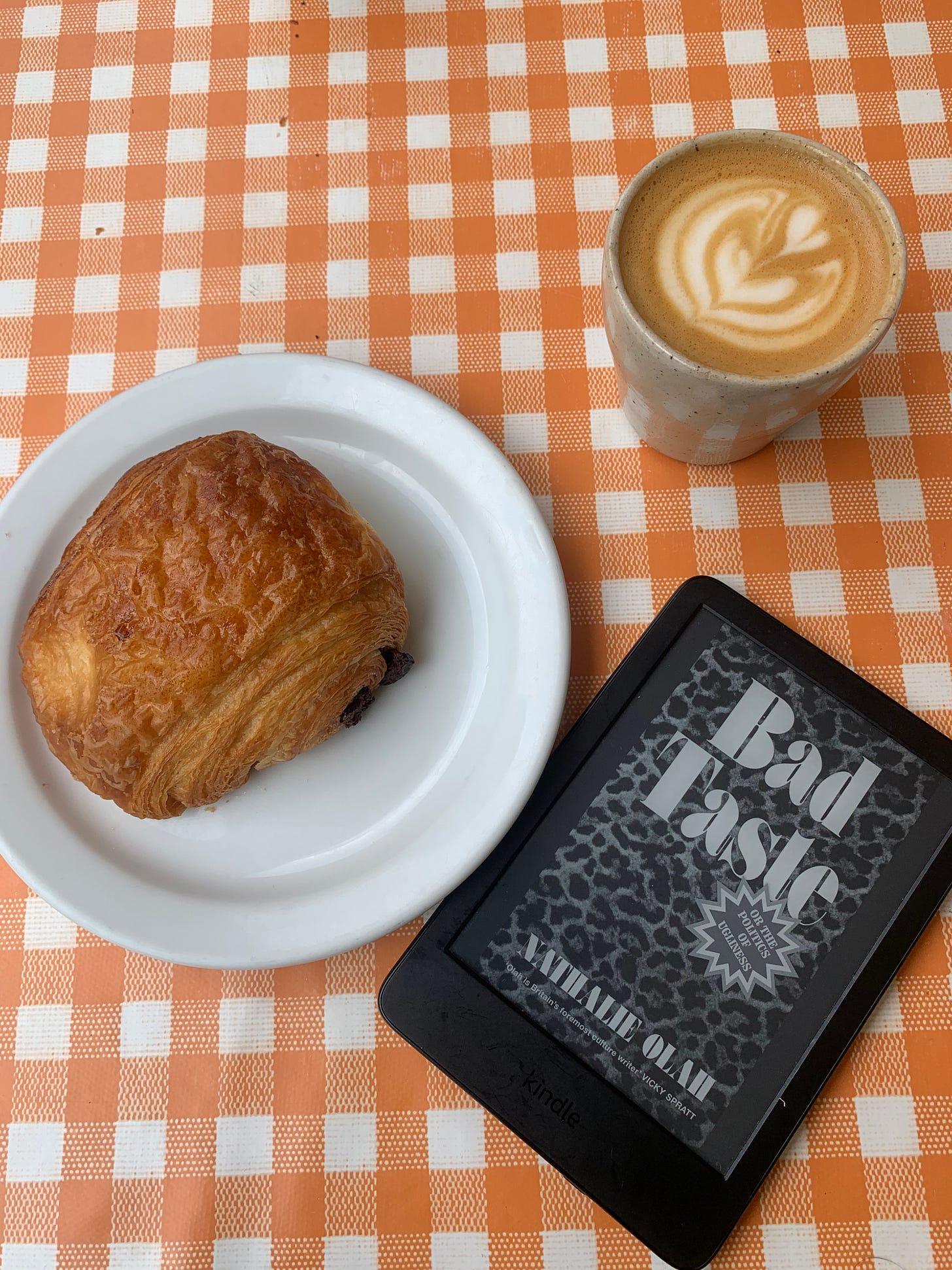 An e-book version of Nathalie Olah's 'Bad Taste' on an e-book reader next to a pain au chocolat pastry on a white plate, and a coffee next to it. All items rest on an orange gingham orange table cover.