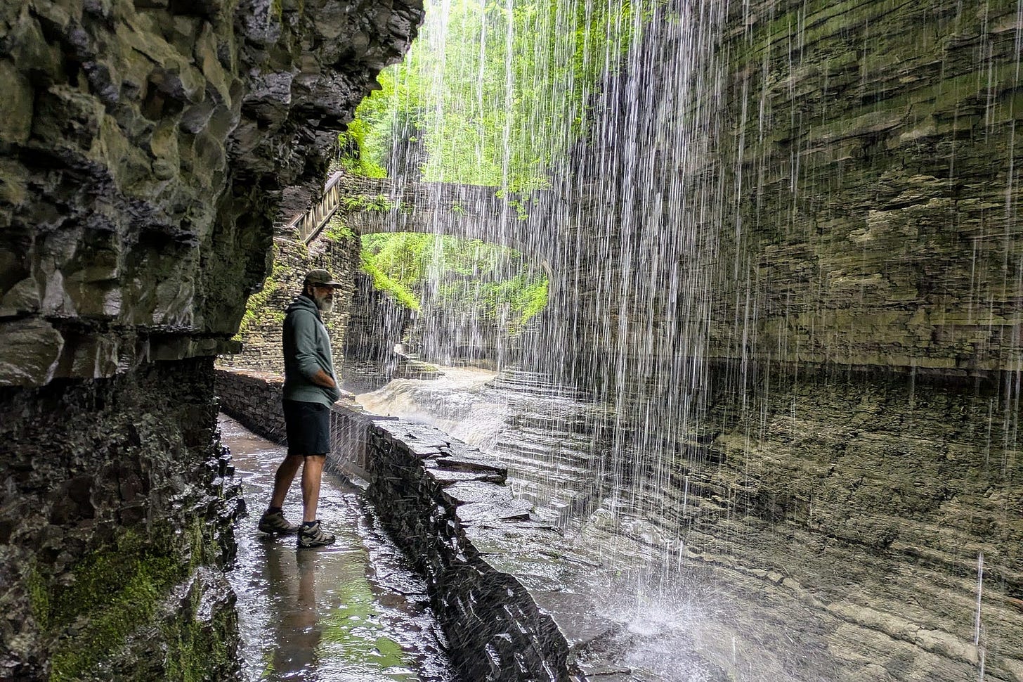 A bearded man in a green long-sleeve shirt, shorts, and hiking shoes stands on a wet stone path behind a cascading waterfall at Watkins Glen State Park. Layered shale rock walls frame the scene, with lush green foliage visible through the curtain of falling water.