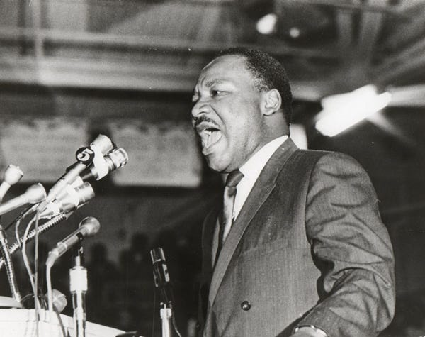 a Black, mustached man in a suit speaks into a podium with a bunch of microphones