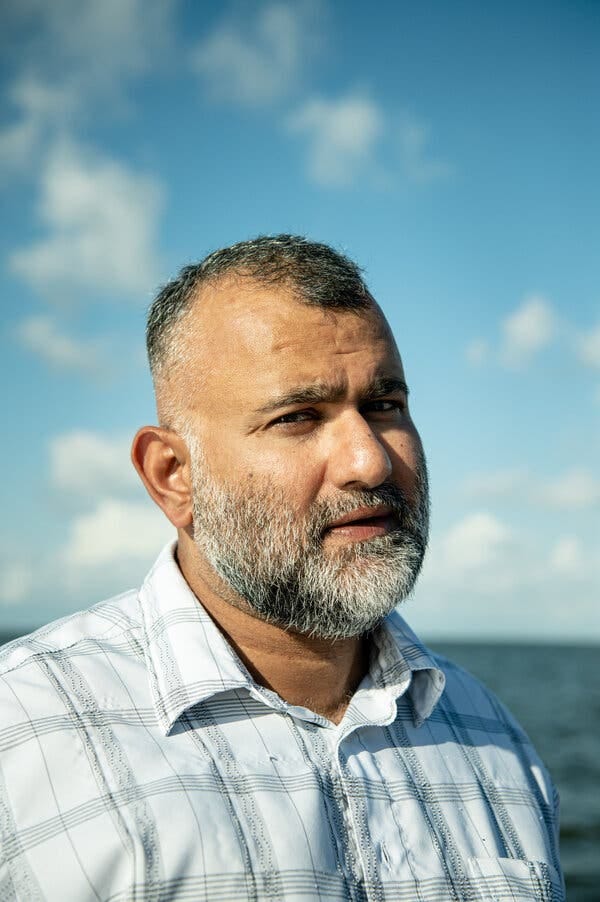 A portrait of Majid Khan with a graying beard wearing a plaid collared shirt in front of the ocean. A portrait of Majid Khan with a graying beard wearing a plaid collared shirt in front of the ocean.