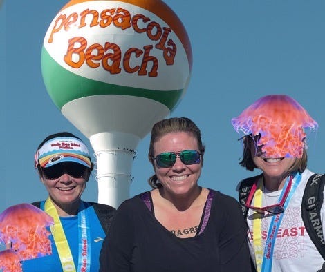 Three women in athletic gear and sunglasses smiling at the camera after a triathlon, with race medals around their necks and decorative jellyfish hats on their heads, with a Pensacola Beach water tower visible in the background.