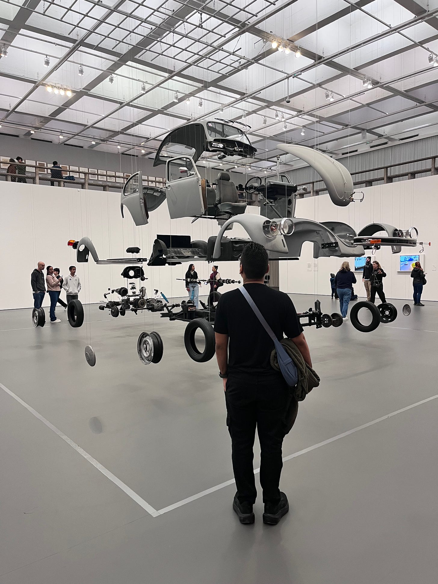 Uma foto vertical no subsolo do Museu de Arte de São Paulo (MASP) em que estou de frente para a instalação artística de Damián Ortega. Na instalação, um fusca está com todos os seus componentes separados, de modo a retratar uma fragmentação.