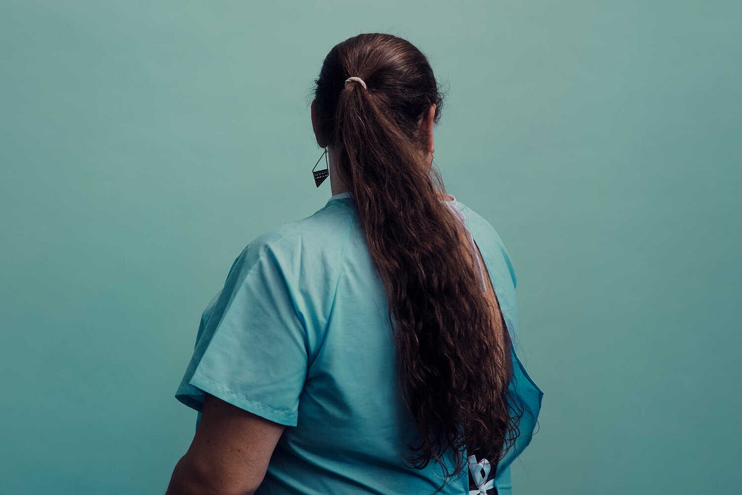 Foto van een forse vrouw met getinte huidskleur en een patiënten operatie jas aan. Ze kijkt weg van de camera, dus je ziet alleen haar lange, donkere krullende haren die in een staart zitten. Je ziet ook een oorbel. De achtergrond is dezelfde turkoois kleur als het kledingstuk.