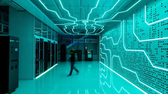 Rows of liquid cooled servers in a brightly lit data centre, with neon circuit patterns on the walls and ceiling. A person walks past the servers