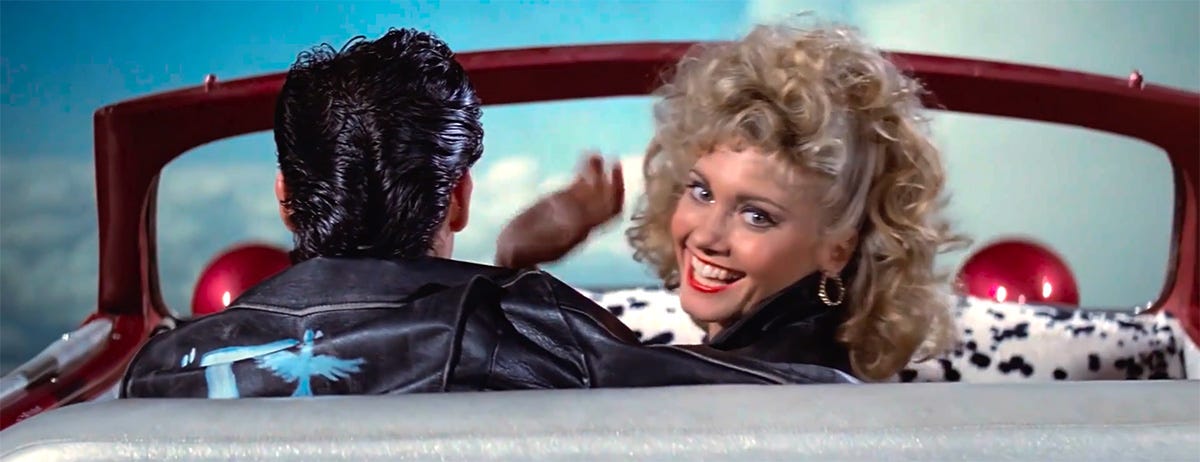 Sandy turns and waves to the camera as Danny pilots Greased Lightning high into the sky - a still from 1978 film Grease