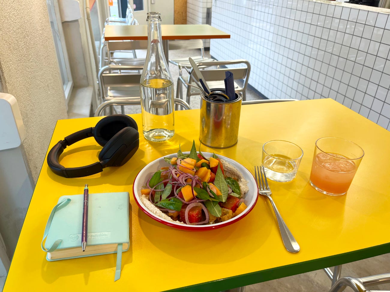 A photo of lunch at a cafe - melon panzanella, grapefruit mimosa, a bottle of water, and headphones next to a small diary on a bright yellow table