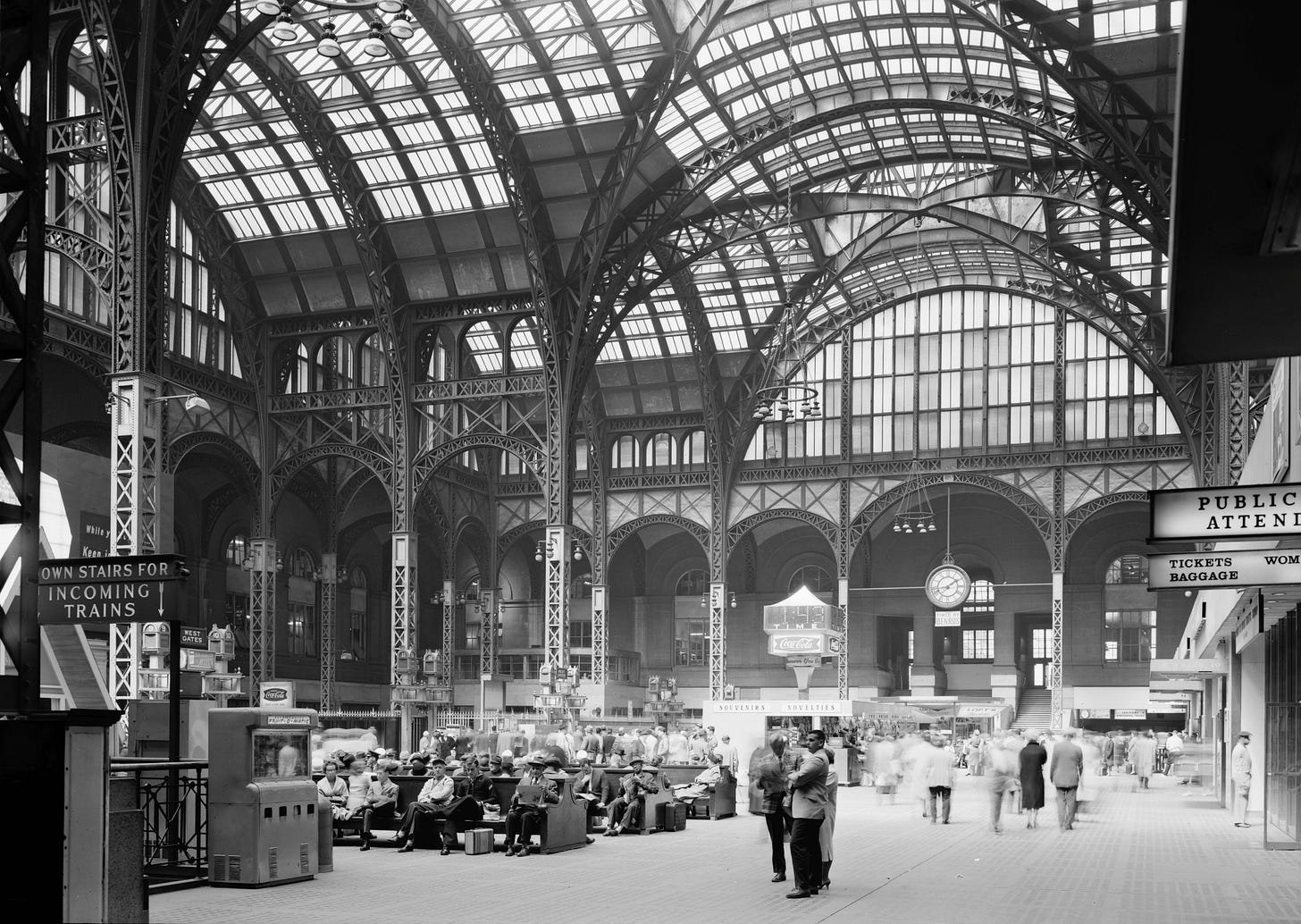 Black-and-white photograph of old Penn Station’s concourse, with passengers and stairways beneath an immense glass-and-steel roof. Black-and-white photograph of old Penn Station’s concourse, with passengers and stairways beneath an immense glass-and-steel roof.