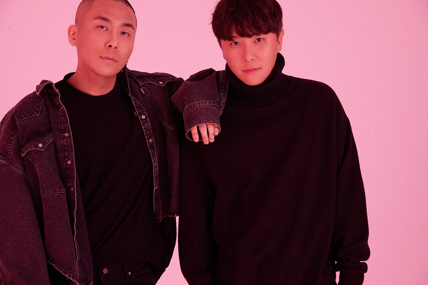 K-pop production duo Black Eyed Pilseung stand in front of a pink background. Rado, on the left, has a buzzcut and is wearing a jean jacket. Choi Kyu-sung, right, has dark hair with bangs and is wearing a turtleneck.