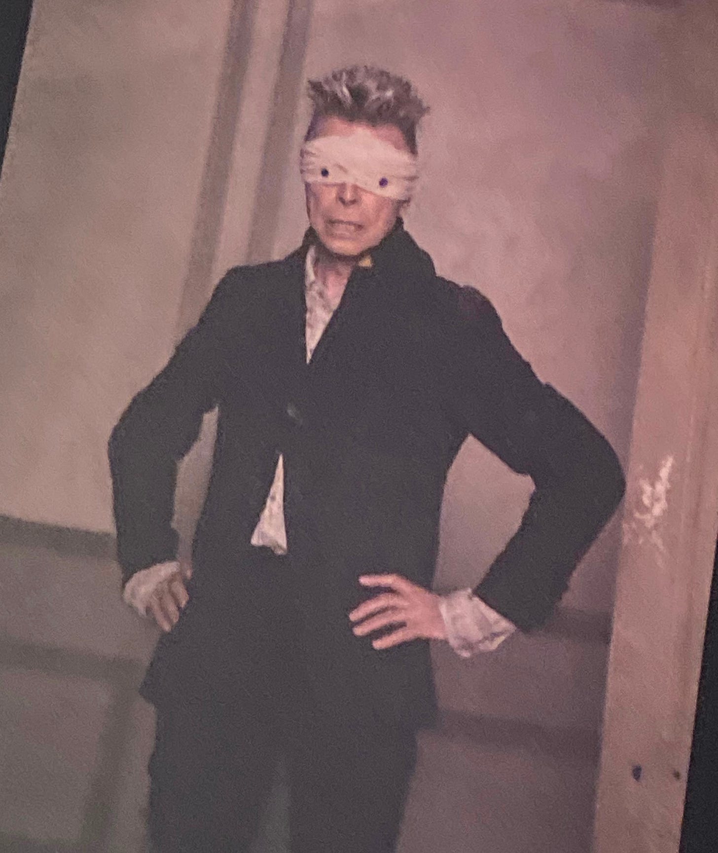 Picture of David Bowie. He is wearing a black suit. His hands are on his hips. He has a blindfold on in which the eyes are cut out. A white curtain appears behind him.