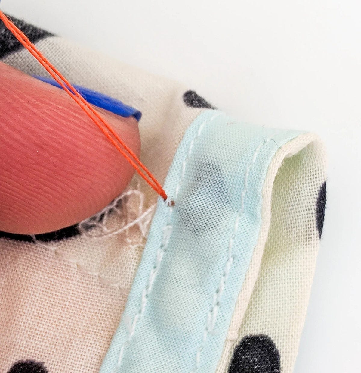 Needle pulling orange thread through fabric near armhole seam with serged edge and light blue binding.