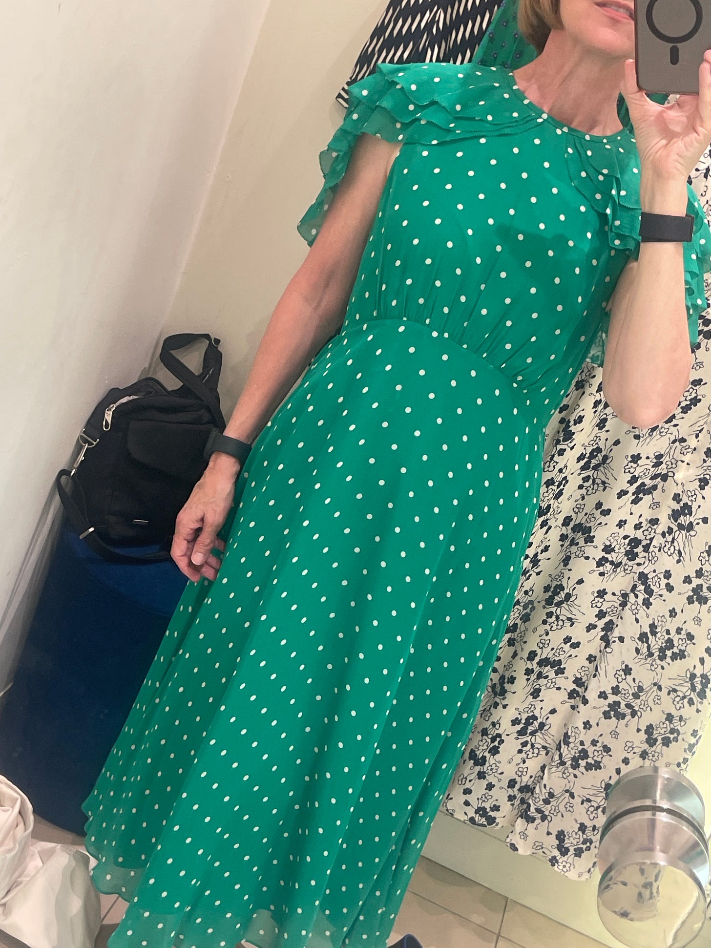 Woman in a fitting room taking a mirror selfie wearing a green polka dot dress with ruffled shoulders and layered hem, with other dresses visible in the background.