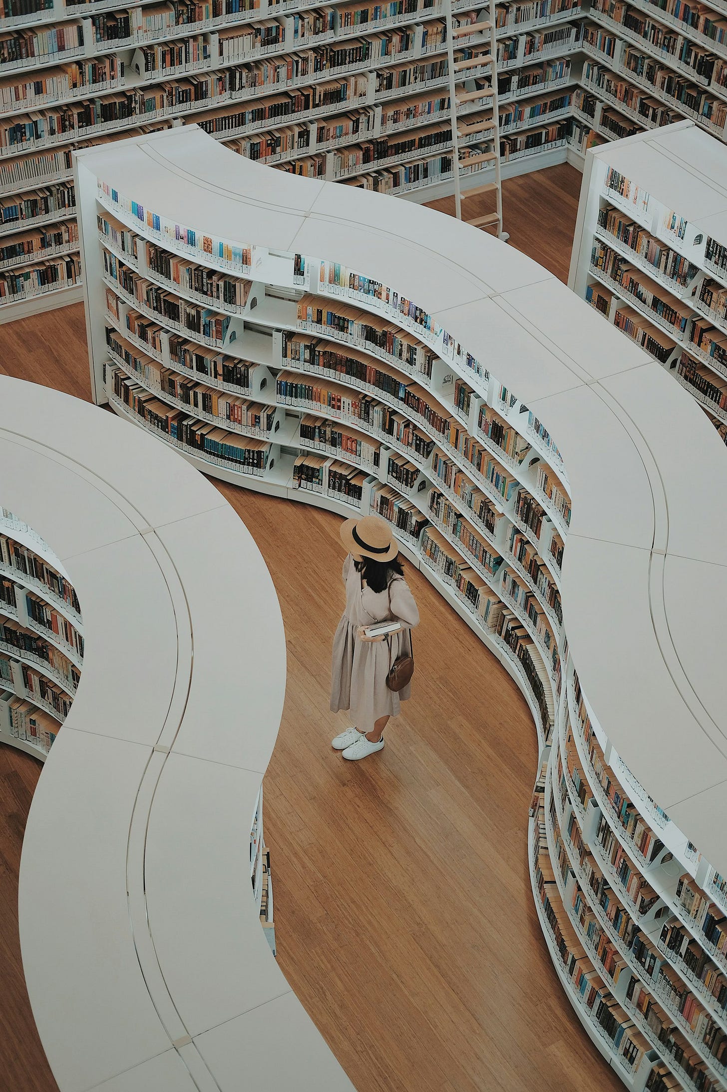 A woman in a tan dress and straw hat standing in a bookstore between two long, winding white bookcases. Many other bookcases surround her.