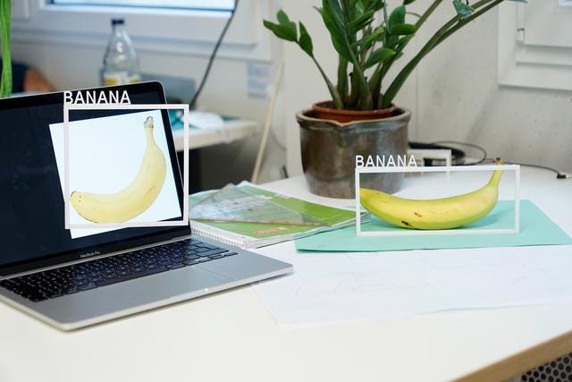 A banana on a table and an image of a banana on a laptop on the same table. Each of the two bananas has a white frame around it with the word ‚Banana‘ sticked on top of it