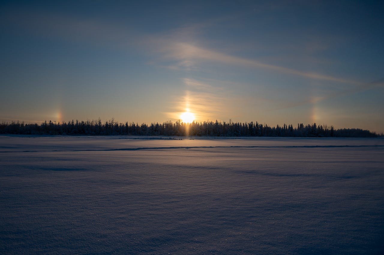 A low winter sun sits just above a dark tree line across a frozen, snow-covered lake. A bright vertical sun pillar extends upward from the sun, and faint rainbow-colored sundogs appear on both sides of the sun against a pale, hazy sky. A low winter sun sits just above a dark tree line across a frozen, snow-covered lake. A bright vertical sun pillar extends upward from the sun, and faint rainbow-colored sundogs appear on both sides of the sun against a pale, hazy sky.
