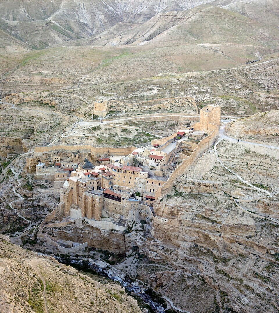 Aerial view of a stone monastery built into the wall of a steep desert canyon, with fortified towers, domes, and terraced buildings.